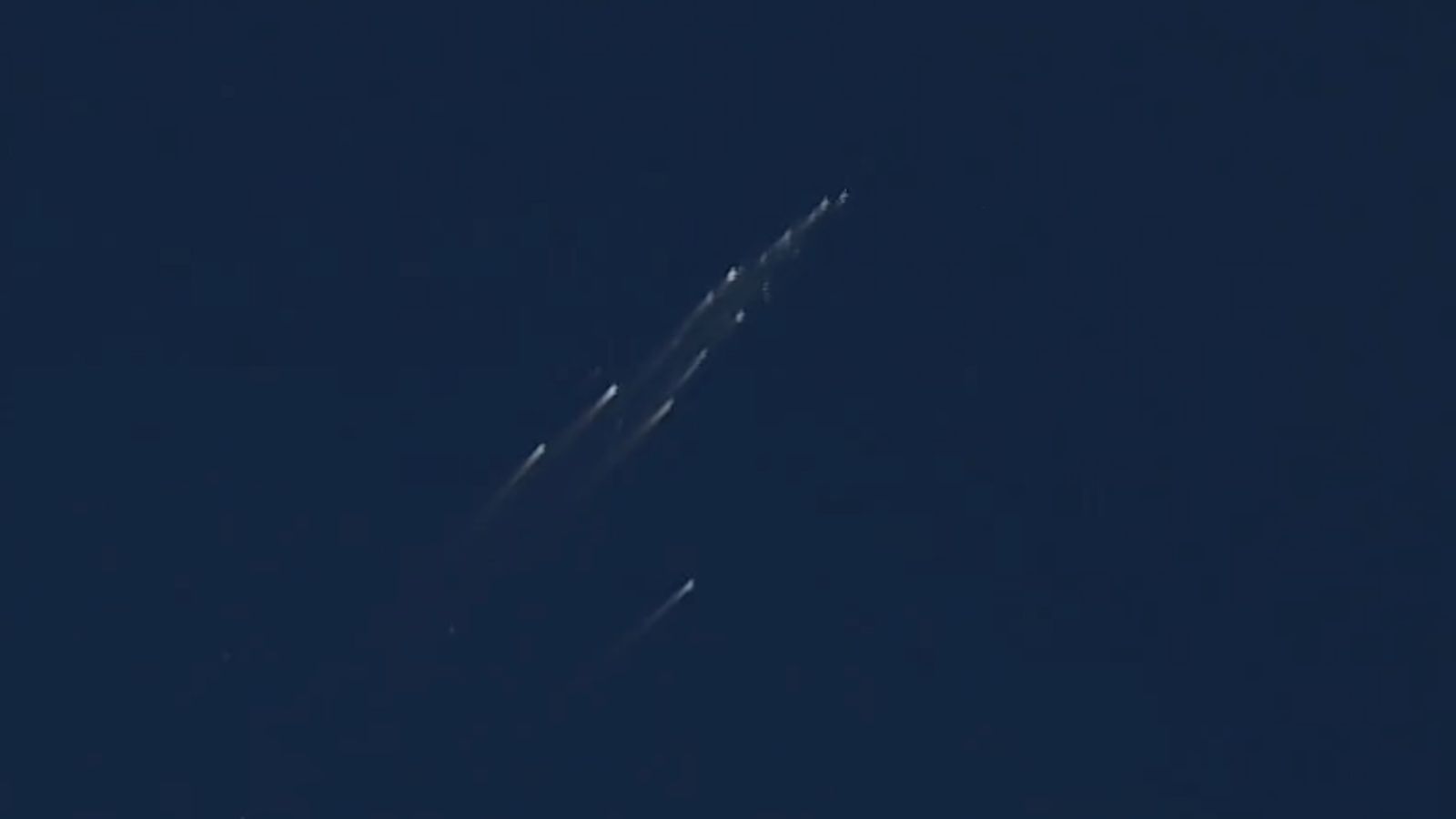 view of a satellite breaking apart, showing a dozen or so bright streaks in a blue sky