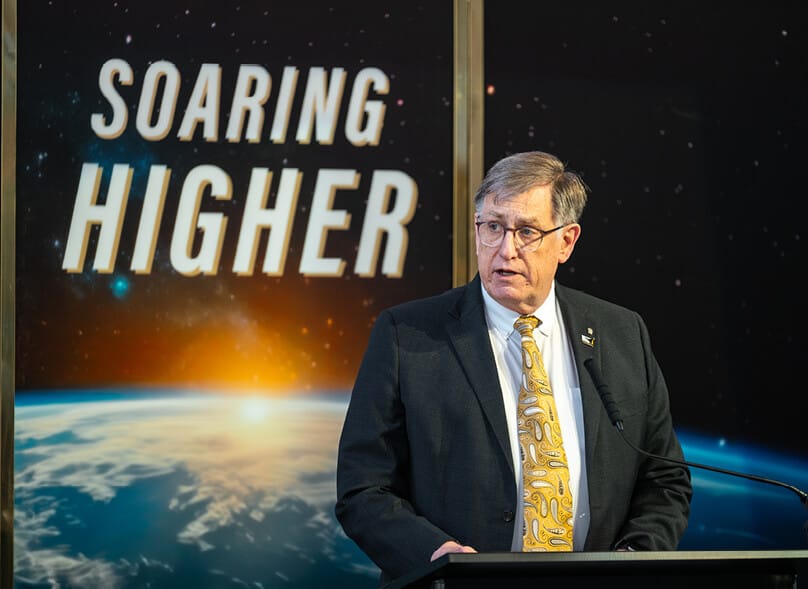 Dan Dumbacher speaks at a podium in front of a backdrop that says “Soaring Higher”