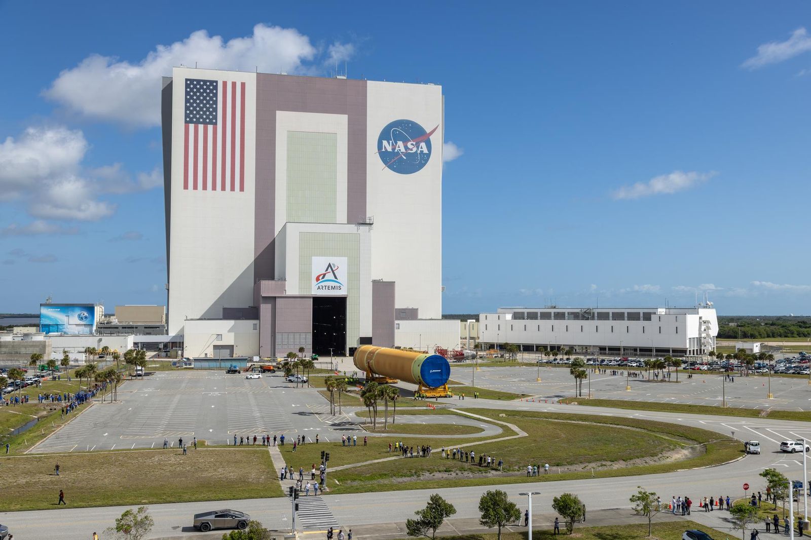 The image shows NASA's Kennedy Space Center iconic Vehicle Assembly Building with an American flag painted on the building along with the NASA and Artemis insignias in a blue sky with some clouds on Tuesday, April 28, 2026.  People gathered to watch the transport of the Artemis III core stage, an orange yellow that will launch during the Artemis III mission. White buildings, grass, and a parking lot can also be seen in the photo. Photo credit: NASA/Glenn Benson