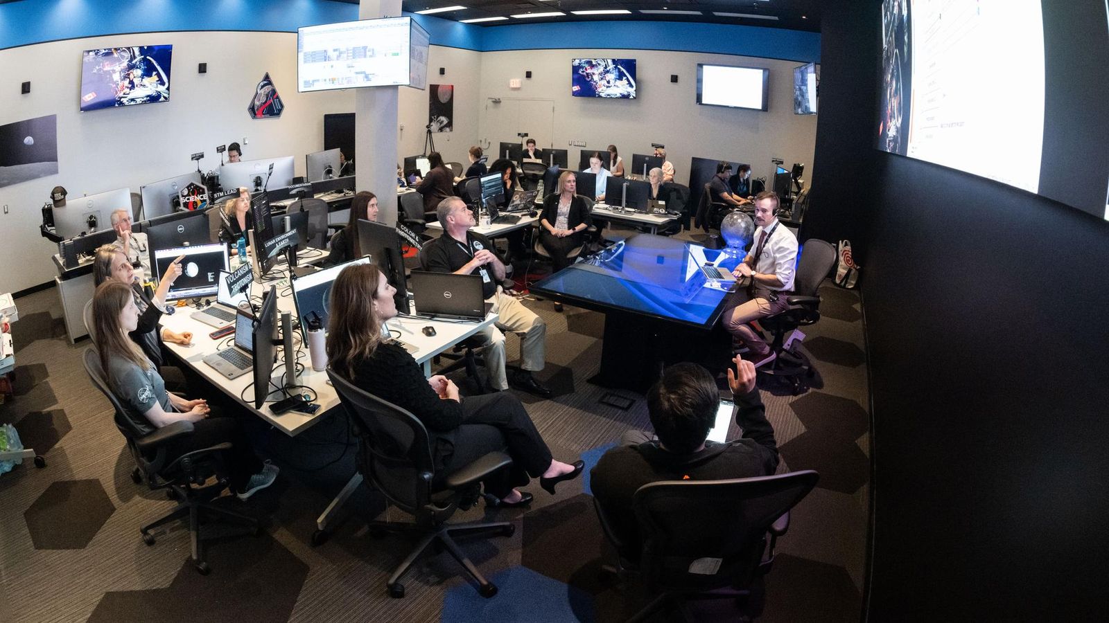 A large operations room filled with about two dozen staff members seated at computer workstations. Rows of desks with multiple monitors face a central area where a person sitting at a glass-topped desk appears to be speaking to the group. Large screens mounted on the walls display space-related imagery and data. Several people are looking towards a large screen hanging on a wall on the right of the image, while others work at their computers. The room is brightly lit, with blue accents along the