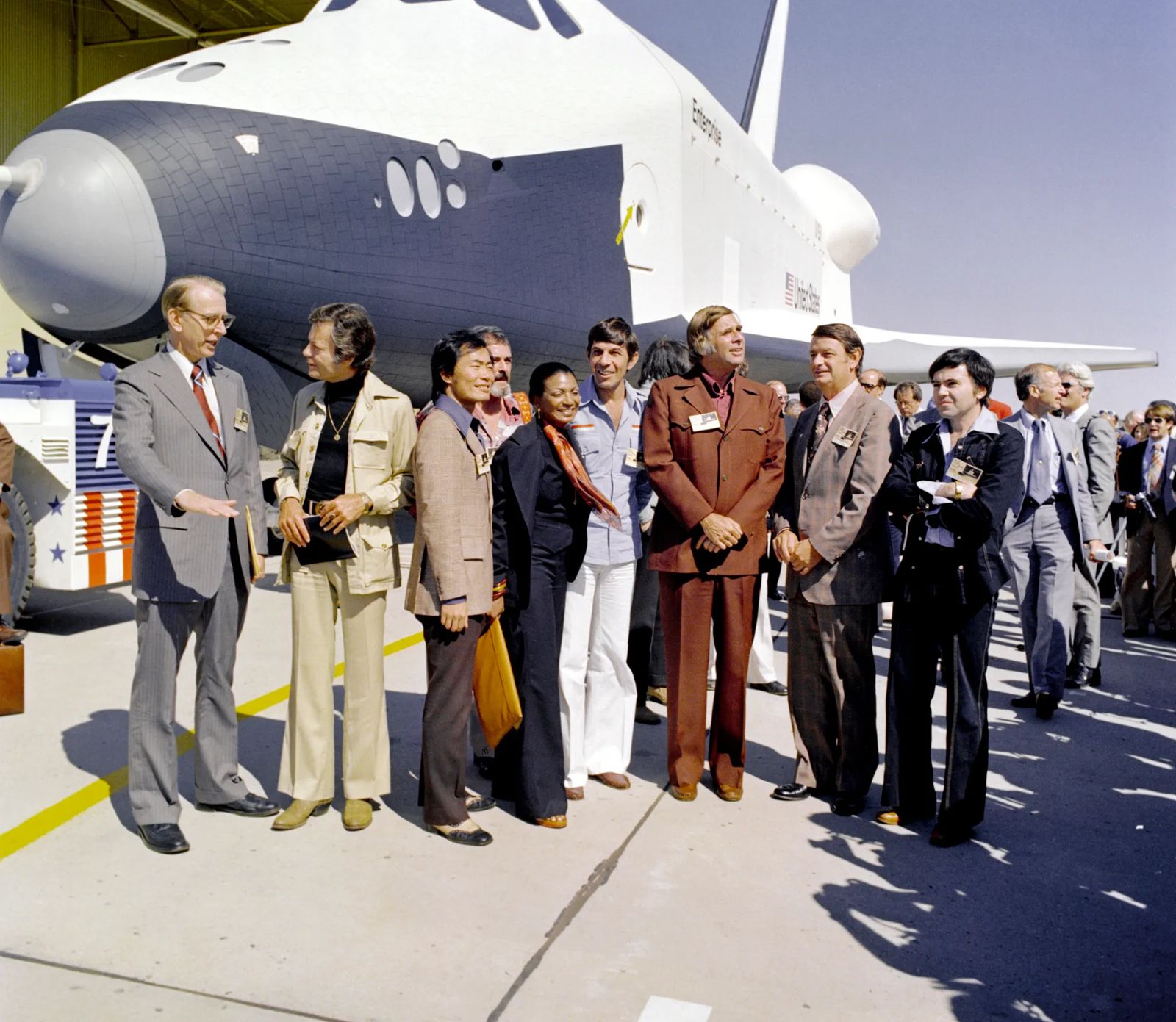 The Shuttle Enterprise (Orbiter Vehicle 101) rolls out of the Palmdale manufacturing facilities with Star Trek television cast members. From left to right they are: Dr. James D. Fletcher, NASA Administrator, DeForest Kelley (Dr. "Bones" McCoy), George Takei (Mr. Sulu), James Doohan (Mr. Scott), Nichelle Nichols (Lt. Uhura), Leonard Nimoy (the indefatigable Mr. Spock), Gene Rodenberry (The Great Bird of the Galaxy), and Walter Koenig (Ensign Pavel Chekov).