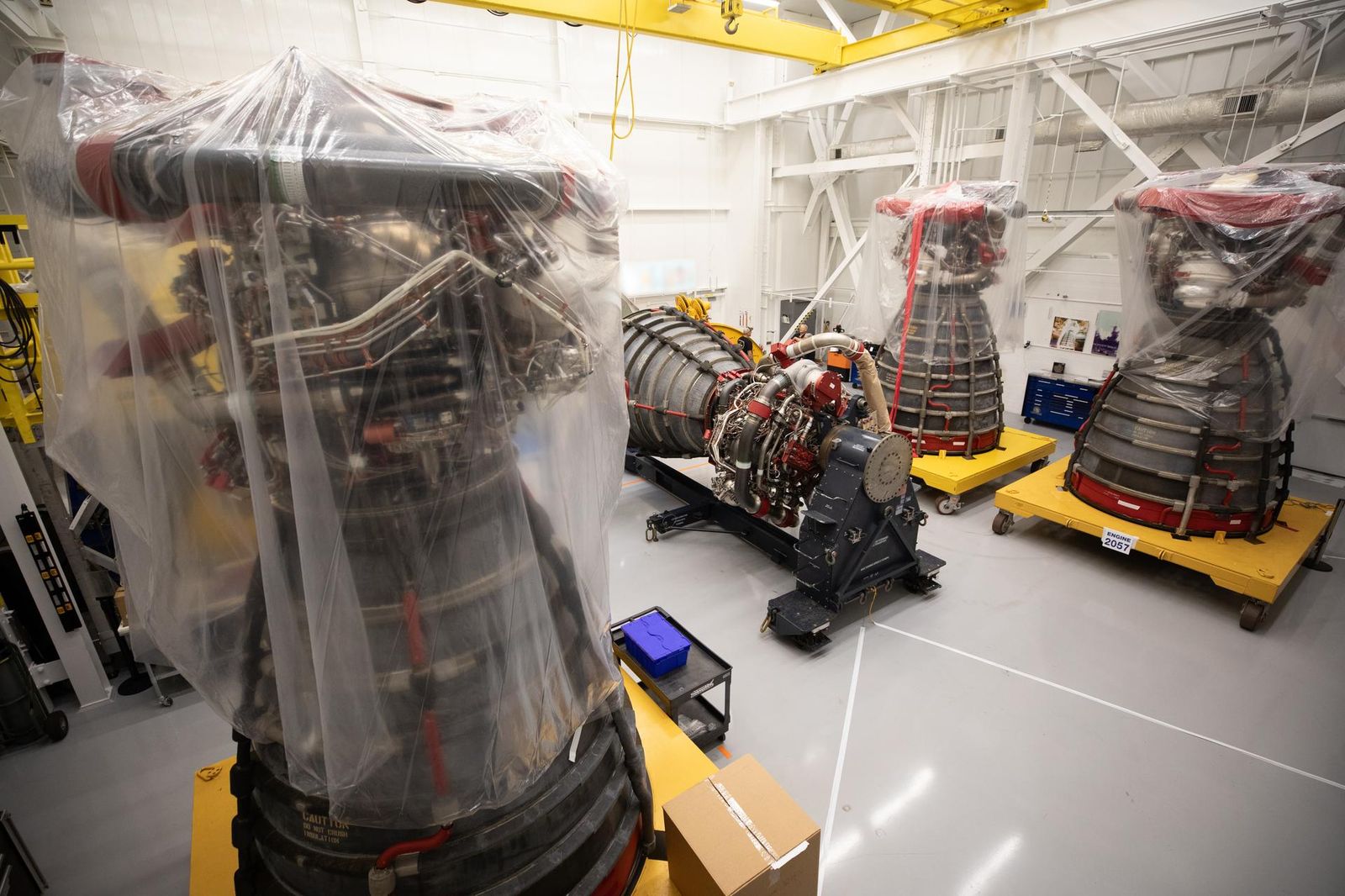 A set of large RS‑25 rocket engines sit inside a bright industrial processing facility at NASA’s Kennedy Space Center. Two engines on the left are wrapped in protective plastic and secured on yellow transport stands, while an unwrapped engine on the right shows detailed plumbing, wiring, and the wide nozzle opening. Overhead cranes, metal platforms, and safety railings surround the workspace, and a technician stands in the background near the wall.