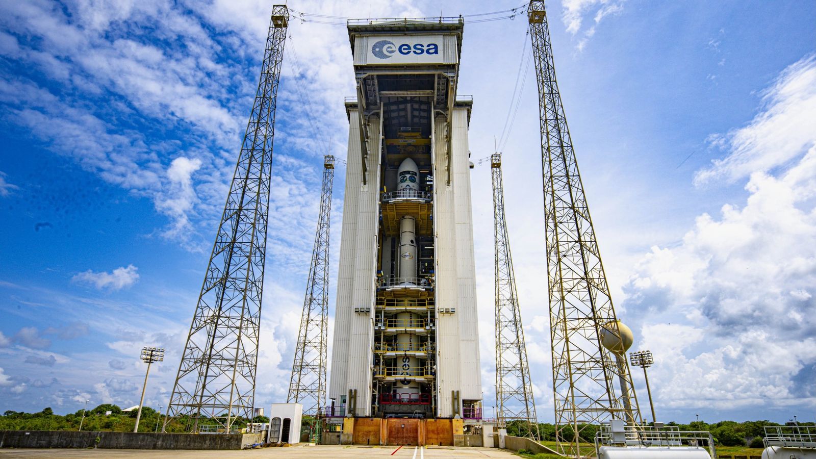 A small white rocket stands vertically inside a white assembly building under cloudy blue skies