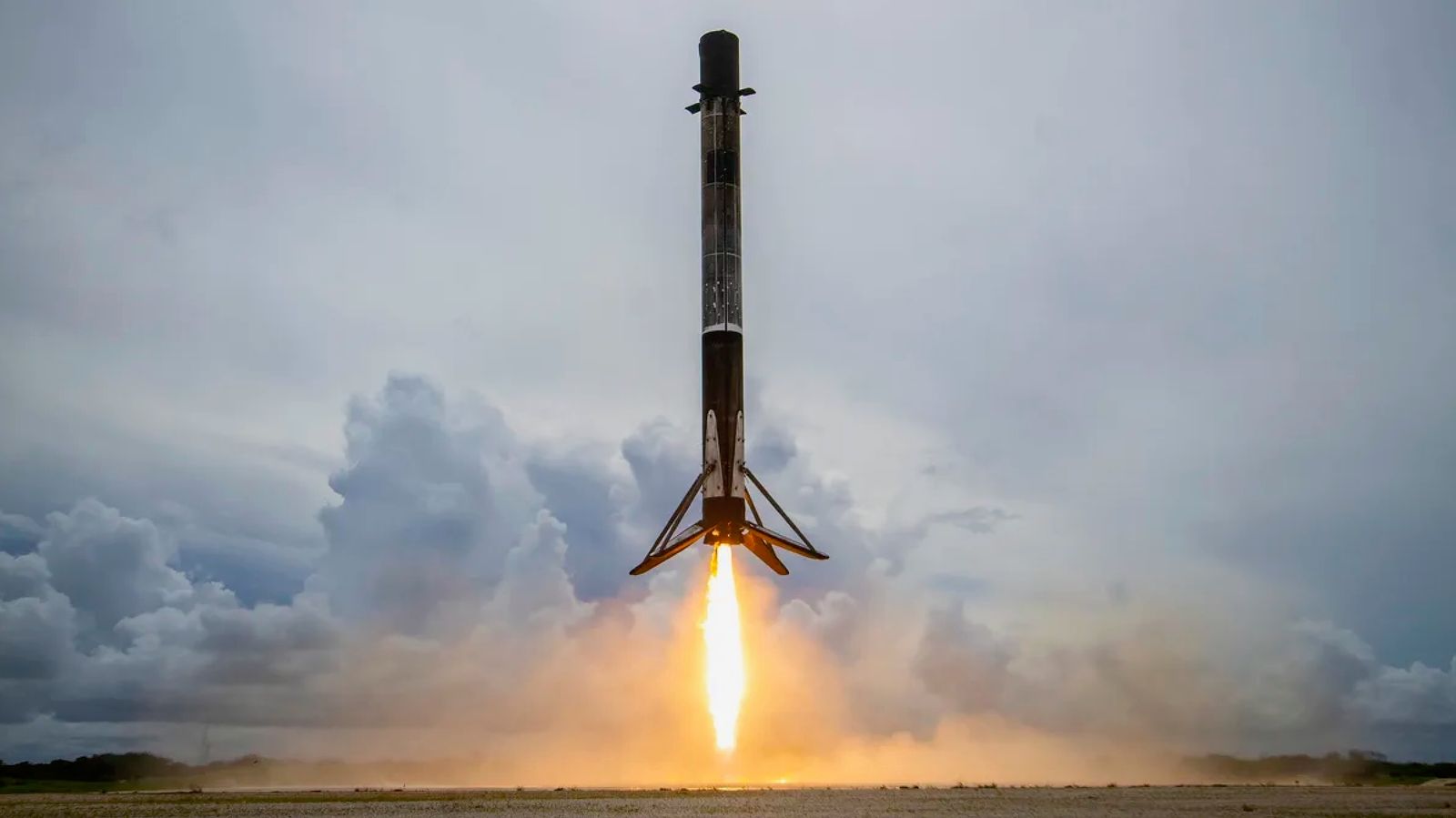 a black and white rocket hovers over a landing pad beneath a cloudy sky