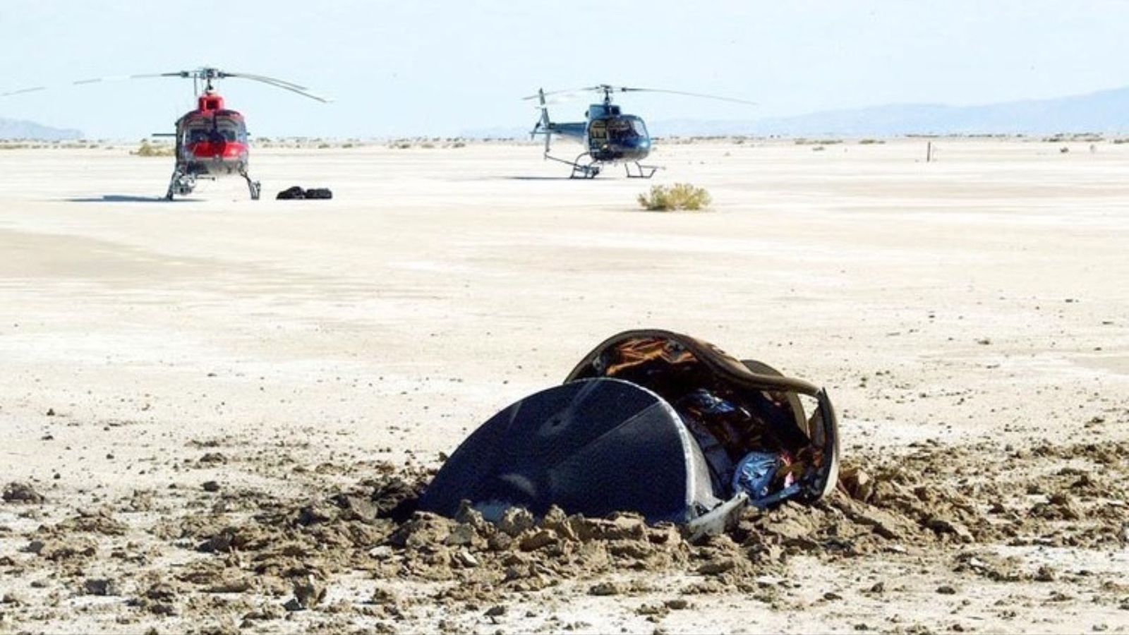 A small pile of metal parts is half buried in a sandy beach as helicopters approach from the background
