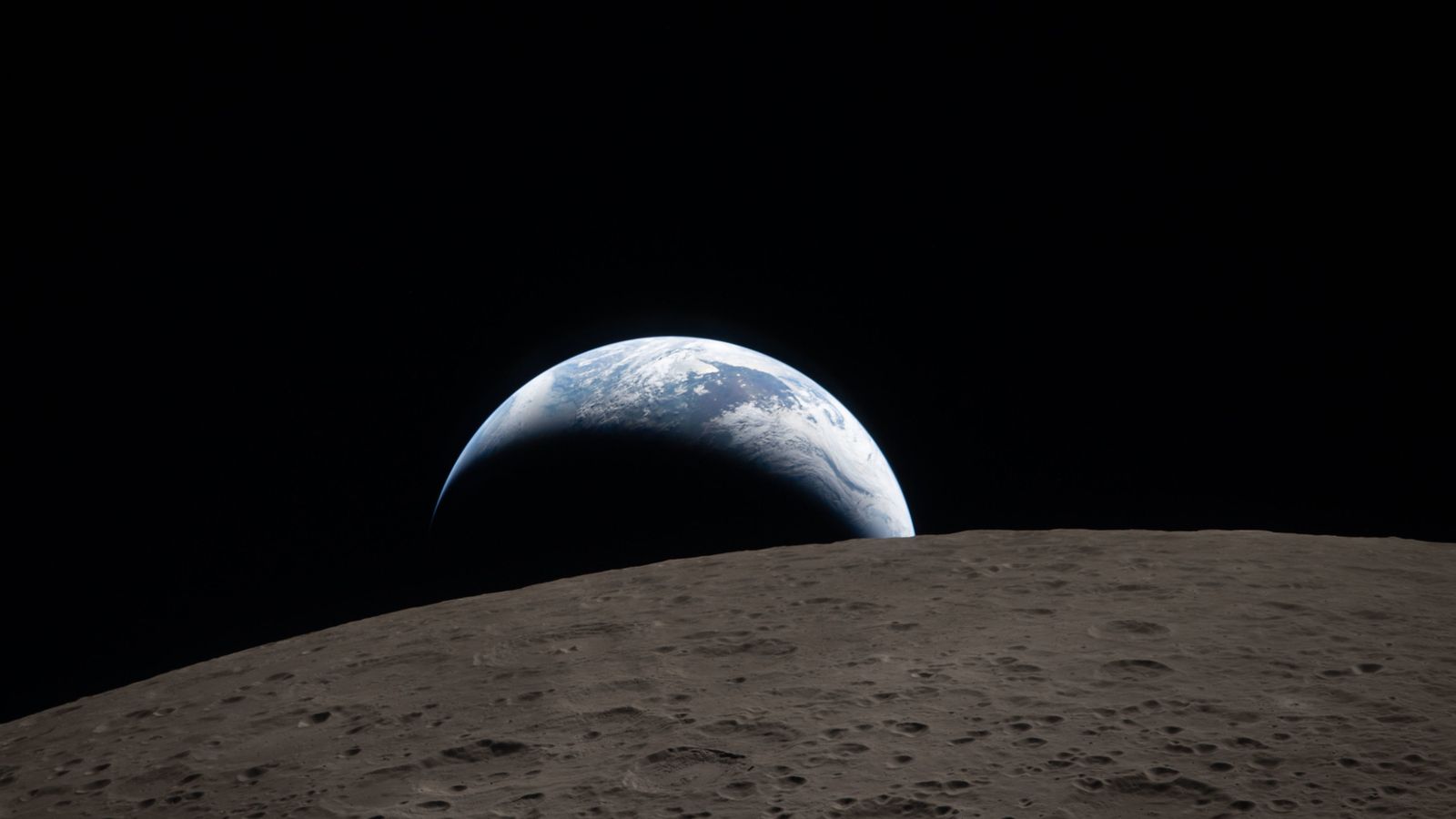 spacecraft photo showing a crescent earth setting above the curved, gray, crater-pocked surface of the moon