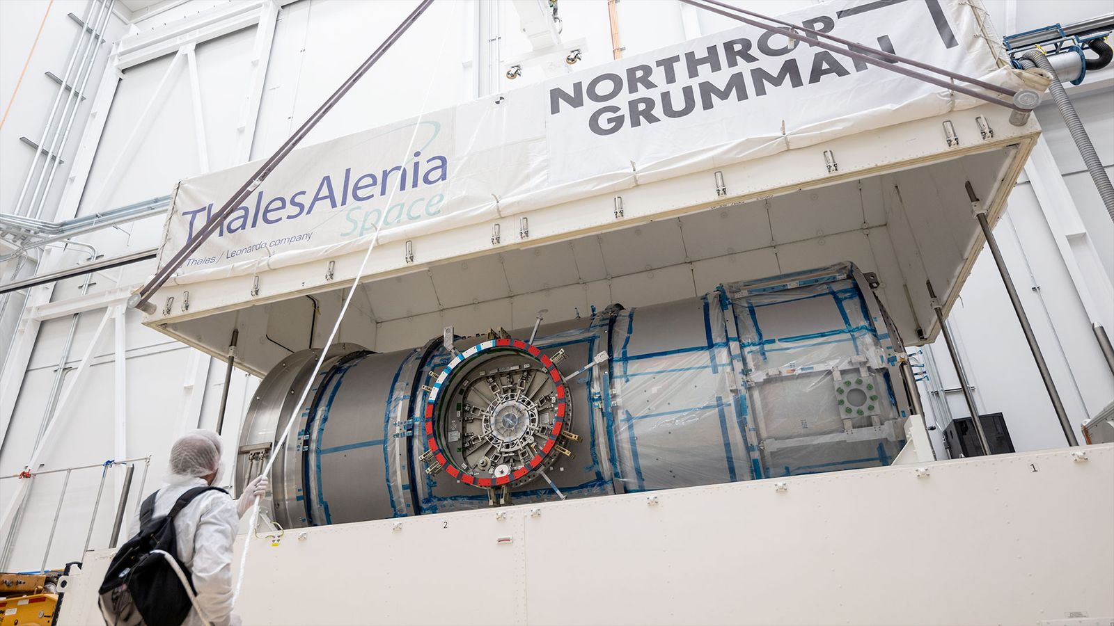 a white top half of a large shipping container is lifted by crane, exposing a metal cylindrical module inside.