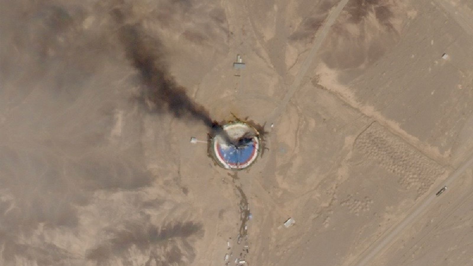 satellite photo of a dark plume of smoke ascending from a blue and red circular launch pad in a desert landscape