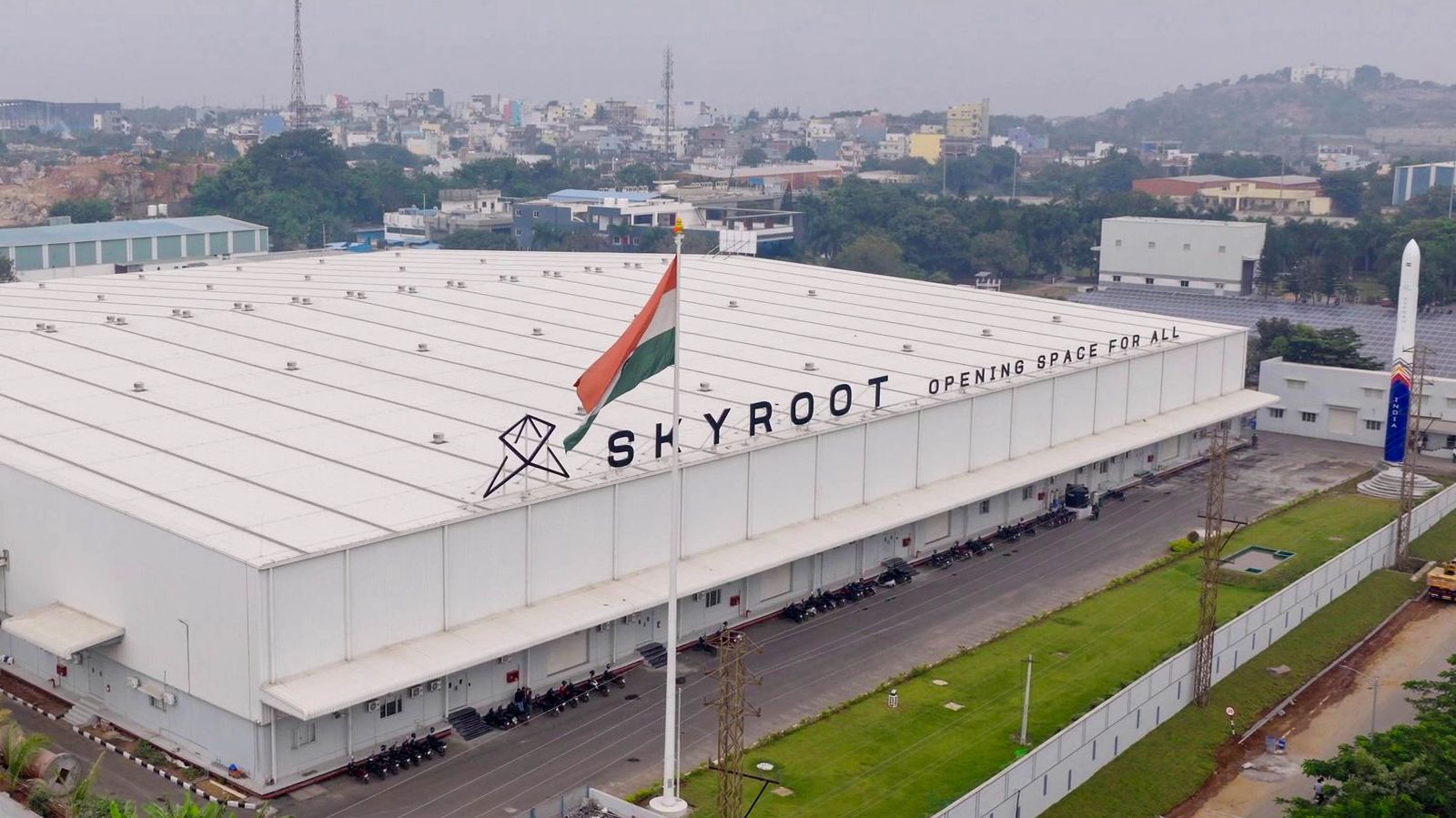 Aerial view of a large, low-slung warehouse-like building with a rocket standing outside it