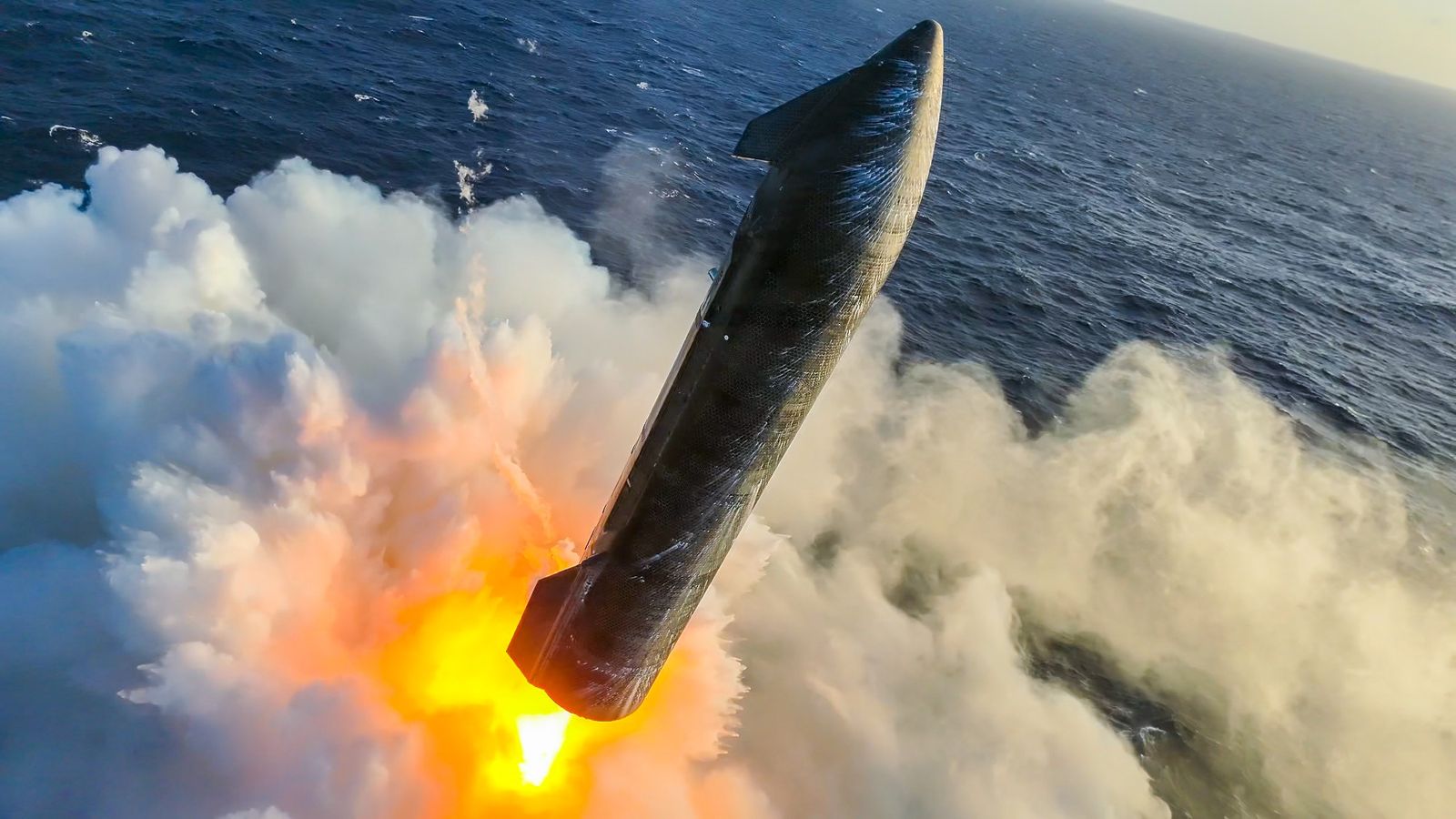 a black rocket with wing-like canards fires its engines just above the ocean, creating a spray of water vapor