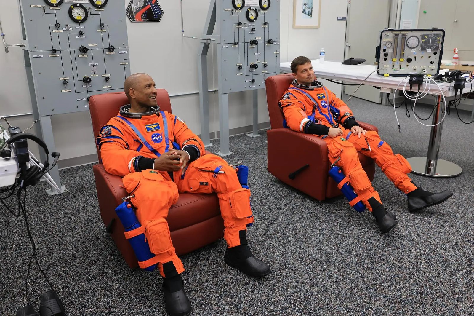 From left, NASA astronauts Victor Glover, Artemis II pilot and Reid Wiseman, Artemis II commander, are seated in chairs wearing orange jumpsuits. Behind them are two large gray panels with dials. The two astronauts are listening attentively and they prepare to conduct leak checks on his spacesuit inside the crew suit-up room in the Neil A. Armstrong Operations and Checkout Building at the agency’s Kennedy Space Center in Florida on Wednesday, April 1, 2026, ahead of the Artemis II test flight.