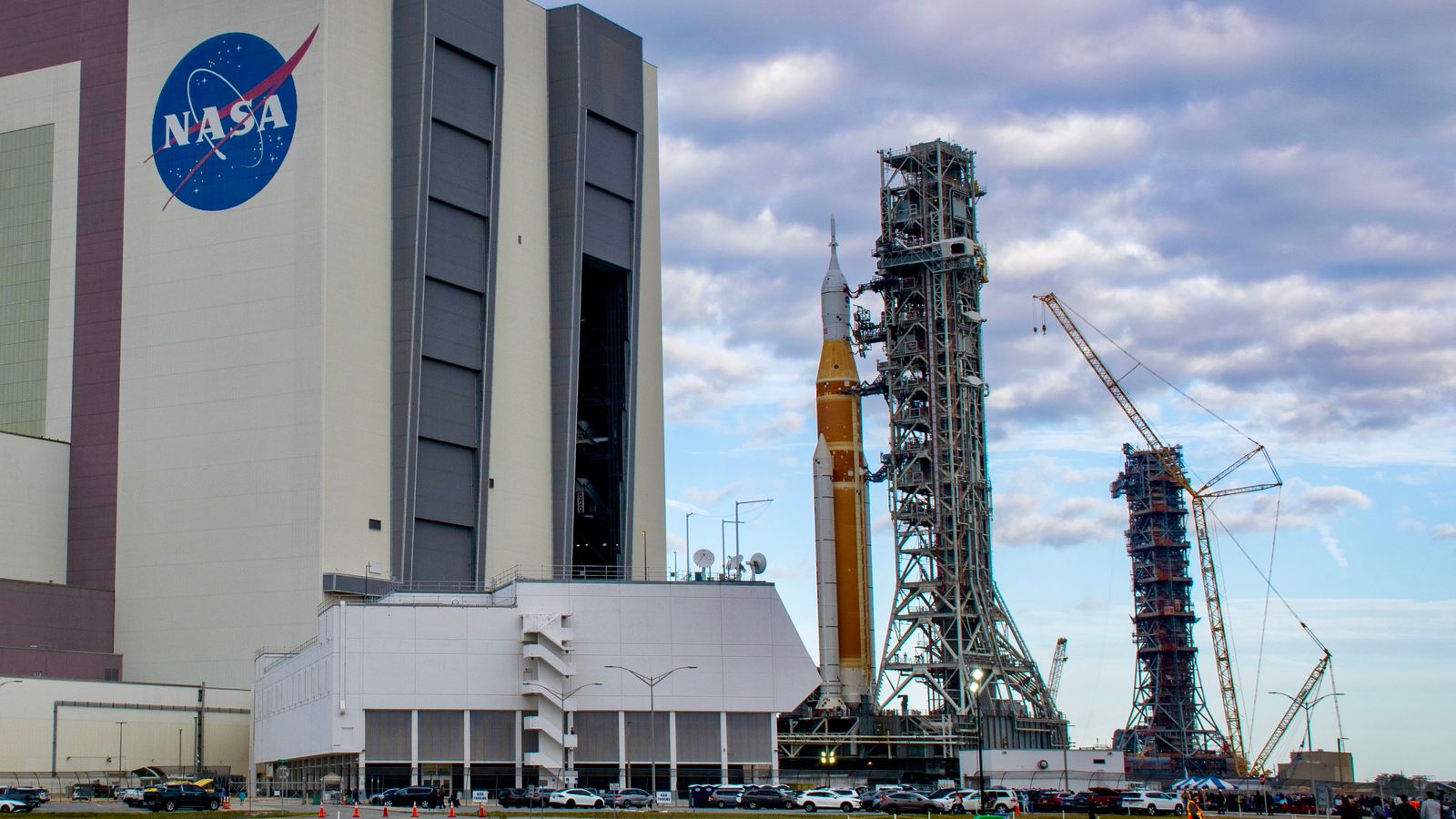 A bit orange rocket rolls out of a giant white building, with an extra launch tower in the background.