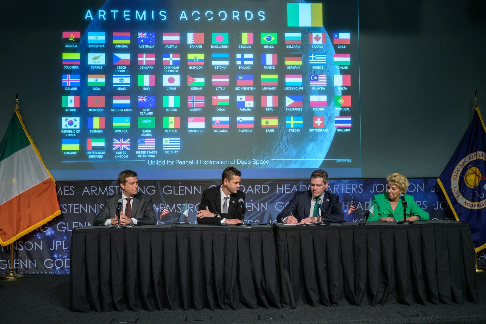 Officials from the United States and Ireland look on as Ireland signs the Artemis Accords while sitting at tables. The flags of the Artemis Accords signatories are behind them.