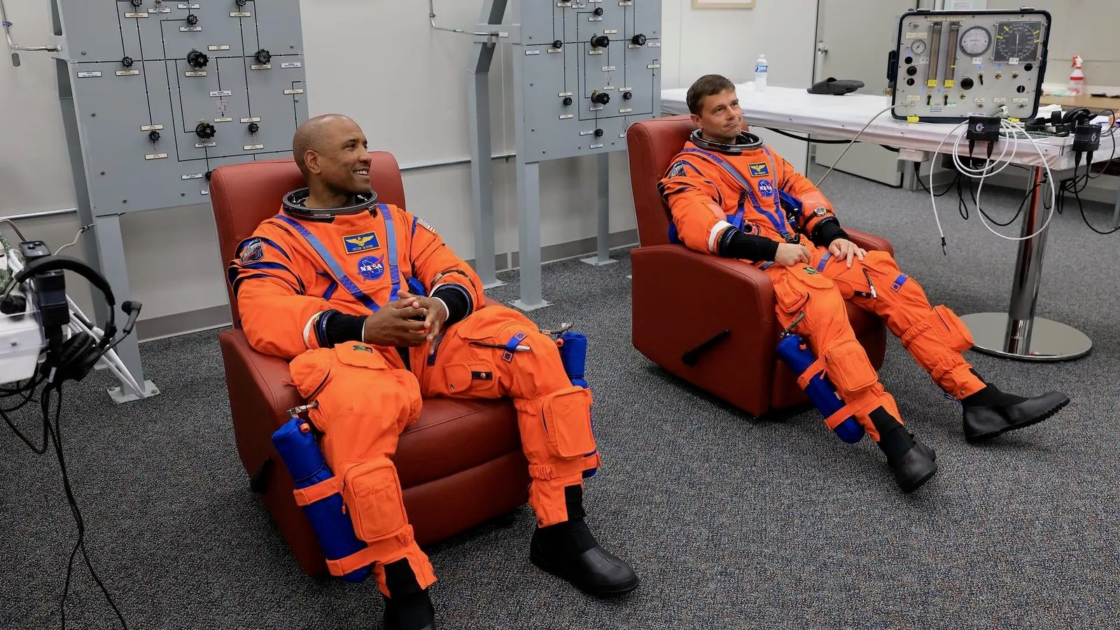 two male astronauts in orange flight suits sit in reddish recliners inside a white-walled room