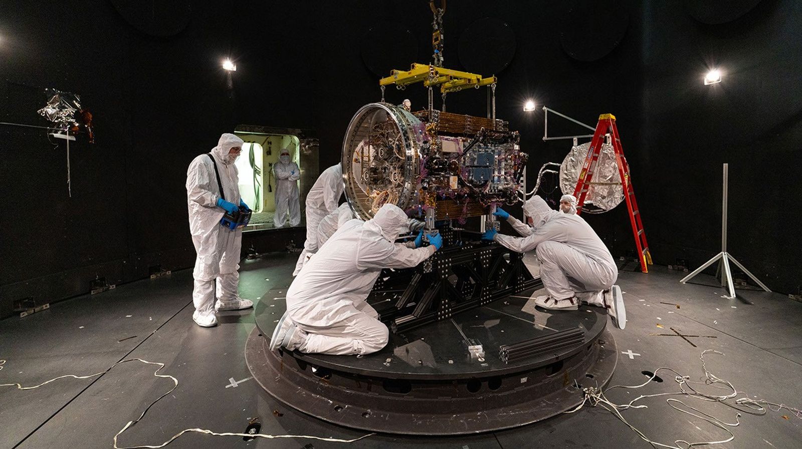 People in clean suits work on a spacecraft in a large chamber.