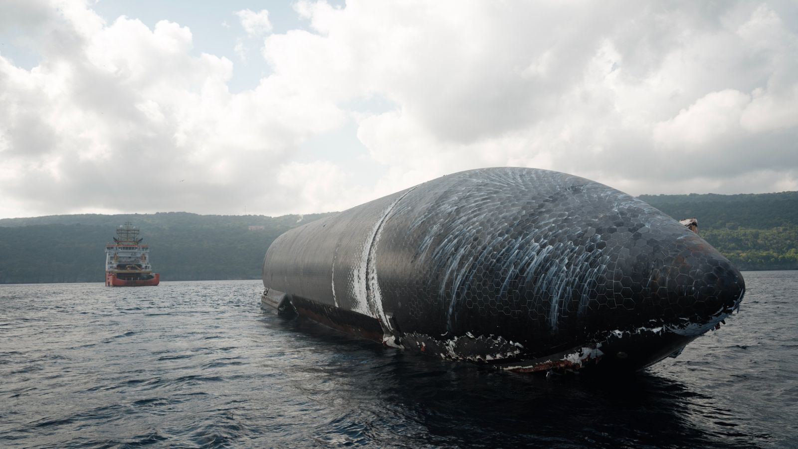 a giant black-tiled rocket lays afloat on the ocean with a boat and an island in the background.