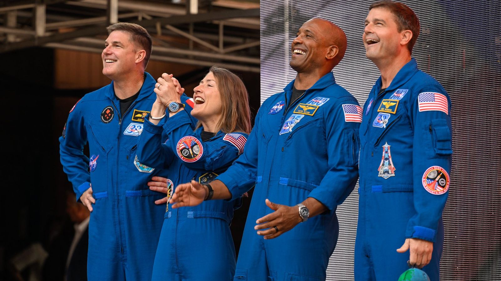 four people in blue flight suits stand on a stage in an amphitheater surrounded by red stone cliffs