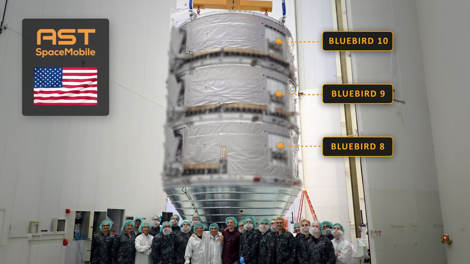 several dozen people stand in front of a stack of three large satellites inside a large, white-walled room