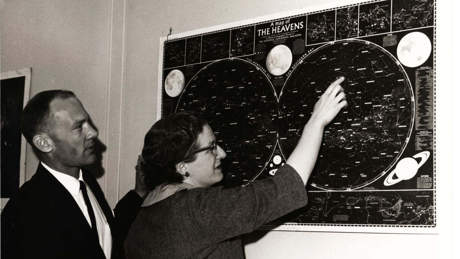 black and white photo of a woman showing a man a wall-mounted star chart