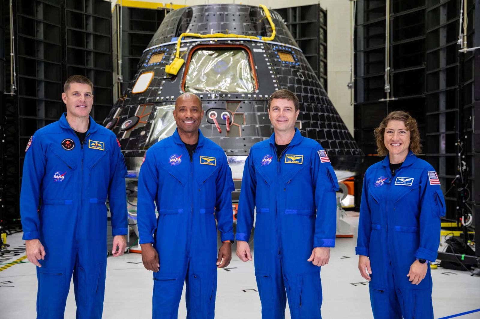 Four people in bright blue jumpsuits pose smiling in front of a spacecraft.