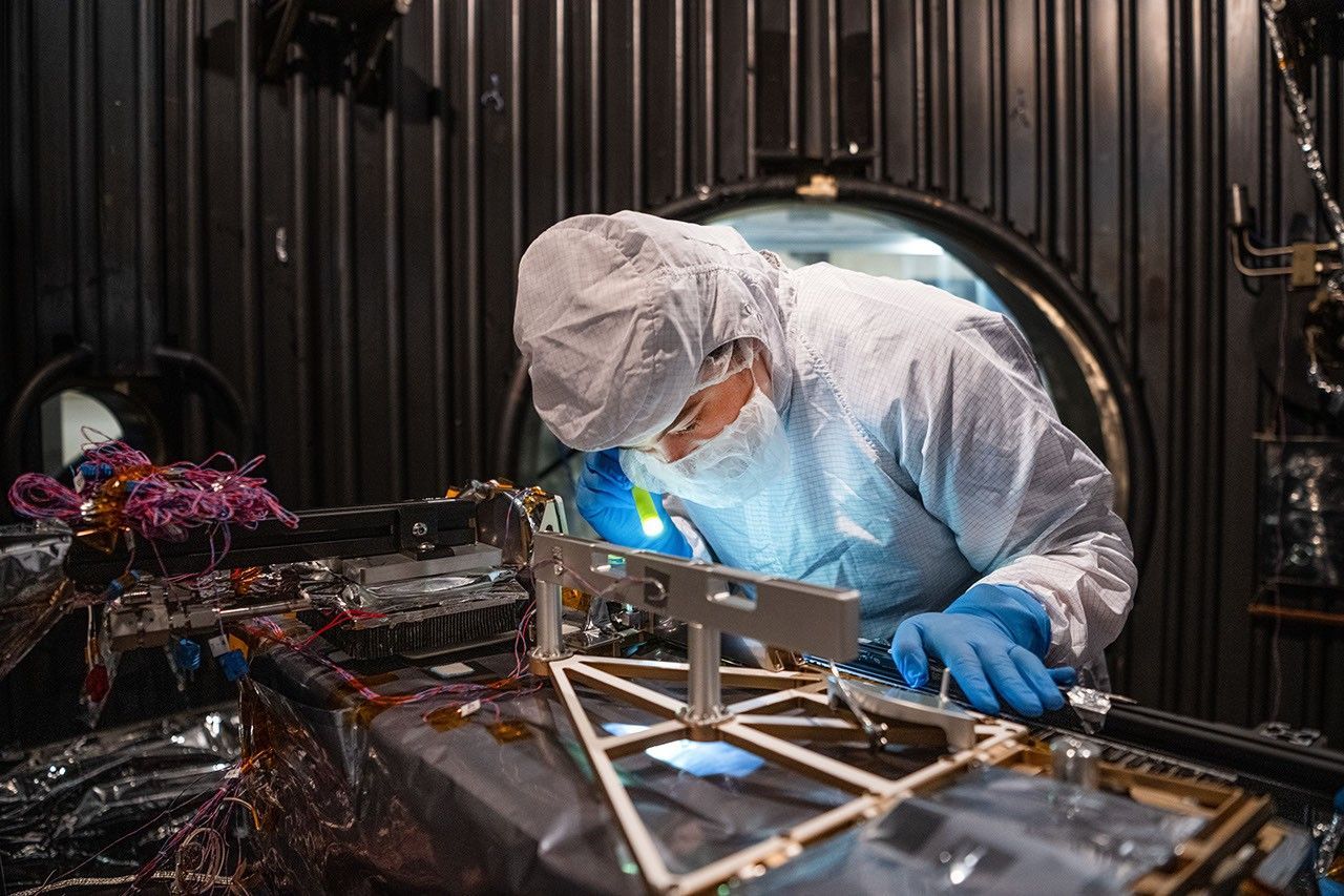 A technician in a white clean-room suit and blue gloves uses a small flashlight to inspect a spacecraft instrument inside a dark testing chamber. Colorful wires and metallic components surround the instrument.