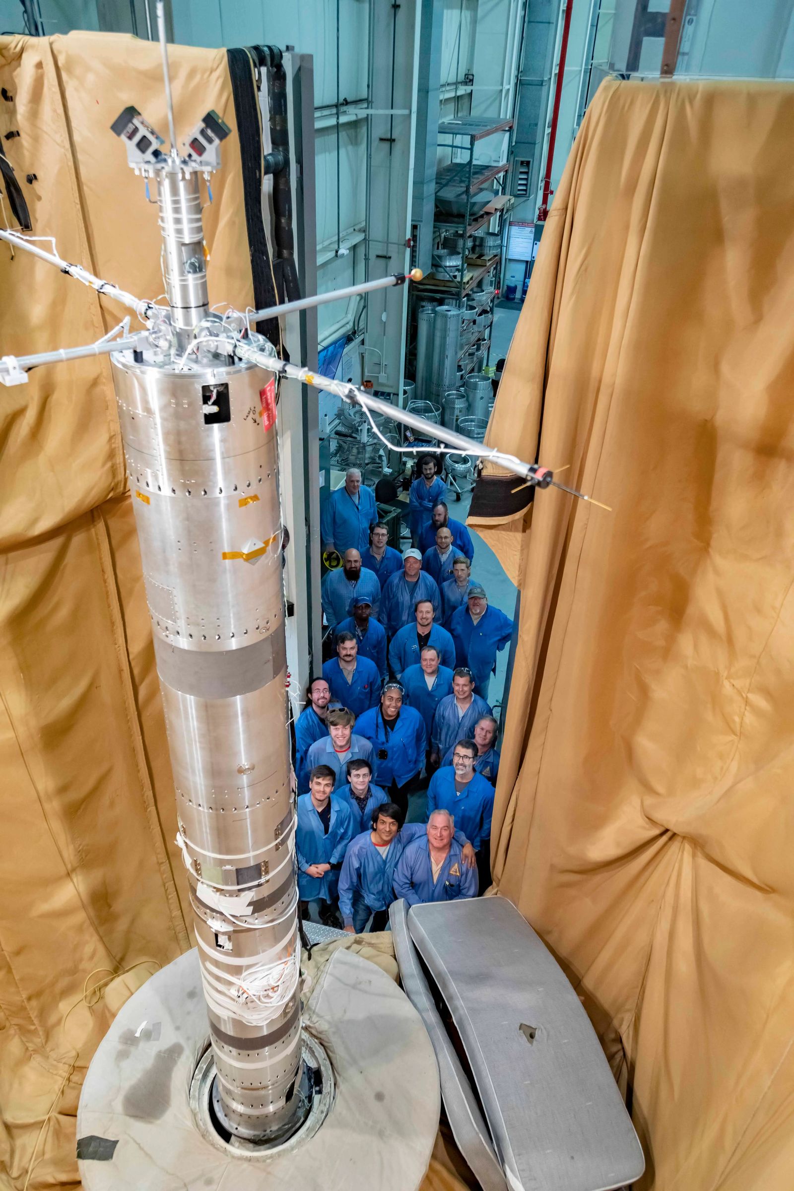 A group of people in blue lab coats stands around a tall, metallic rocket component inside an industrial facility with beige protective curtains.