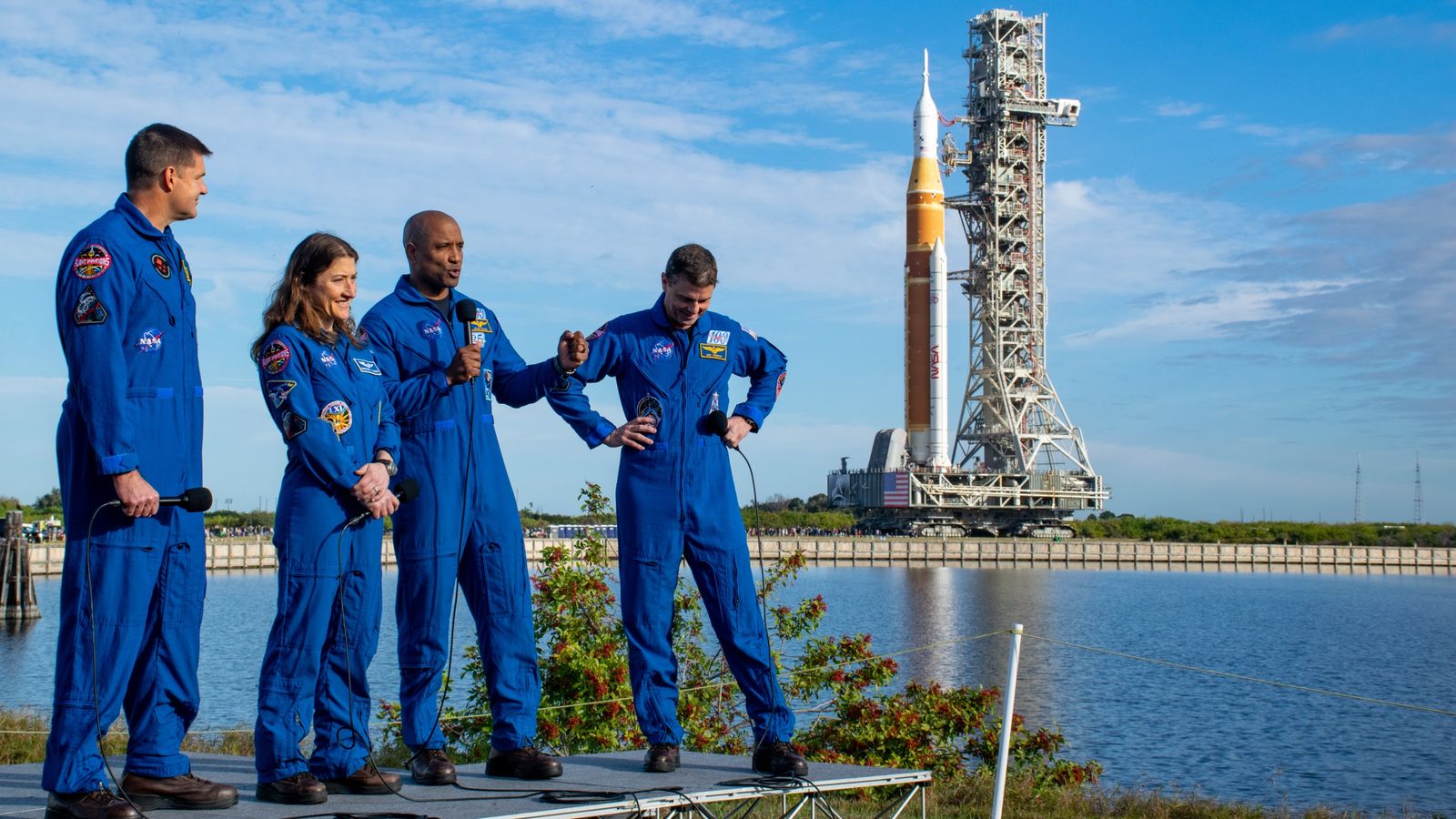 Four humans in blue jumpsuits stand outside on a raised platform. A big orange rocket and launcher tower can be seen in the background.