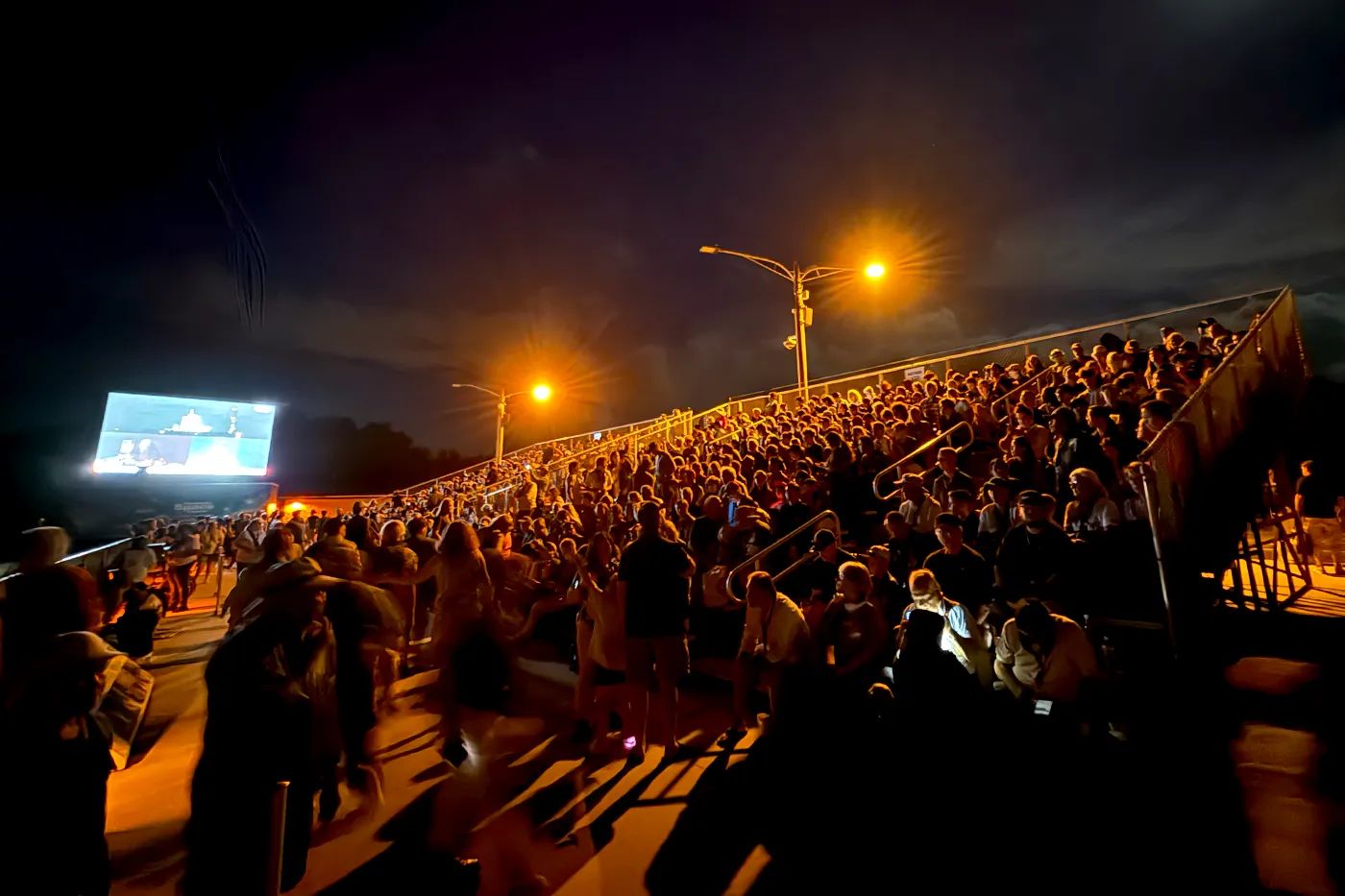 A large crowd gathered along a waterfront at night to watch a launch, with a screen and lights visible in the distance.