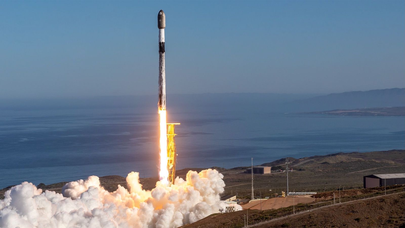 a black and white rocket launches into a blue sky from a seaside pad