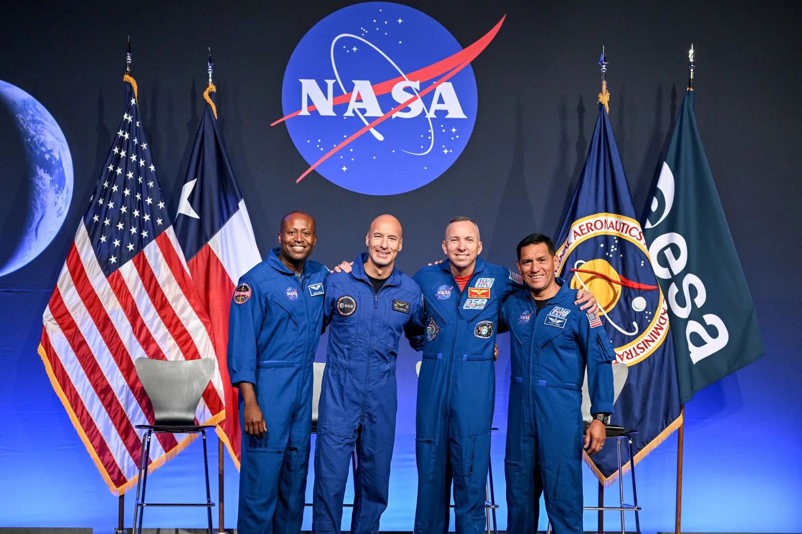 From left to right, NASA astronaut Andre Douglas, ESA (European Space Agency) astronaut Luca Parmitano, and NASA astronauts Randy Bresnik and Frank Rubio take a photo together. Their arms are linked around each other's shoulders. Behind them are the flags of the United States, Texas, NASA, and ESA.