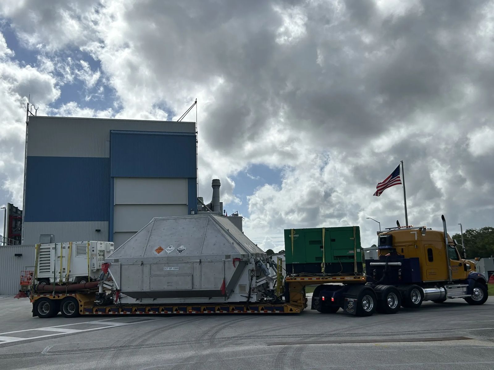 A large truck with a yellow cab hauls a wide, metal, container-like structure on a low trailer, with additional equipment secured behind it. An American flag flies from the truck as it sits on a paved area beside a tall industrial building. Thick, backlit clouds fill the sky above at NASA's Kennedy Space Center in Florida on Tuesday, April 28, 2026. Photo credit: NASA/Tiffany Fairley