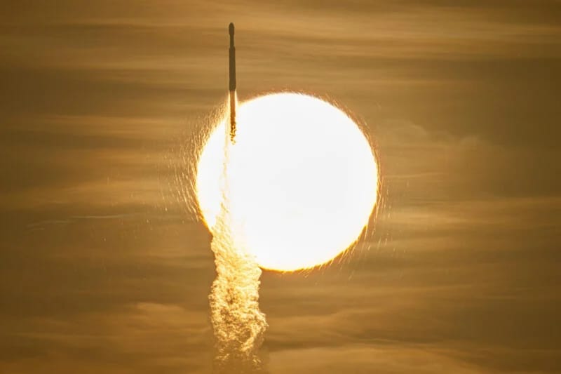 A rocket ascends vertically, silhouetted against the bright, golden disk of the sun in a hazy sky.