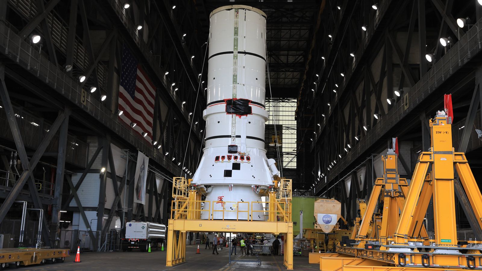 The bottom segment of a white solid rocket booster stands on a yellow support inside a giant rocket hangar.