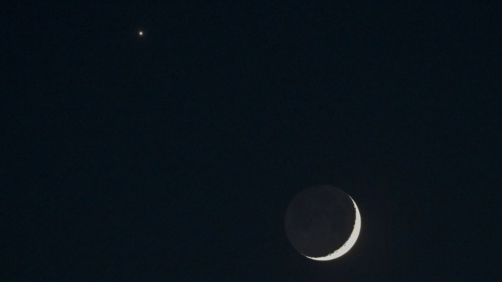A thin crescent moon shines in a black sky with Venus appearing as a bright star-like object to its upper left.