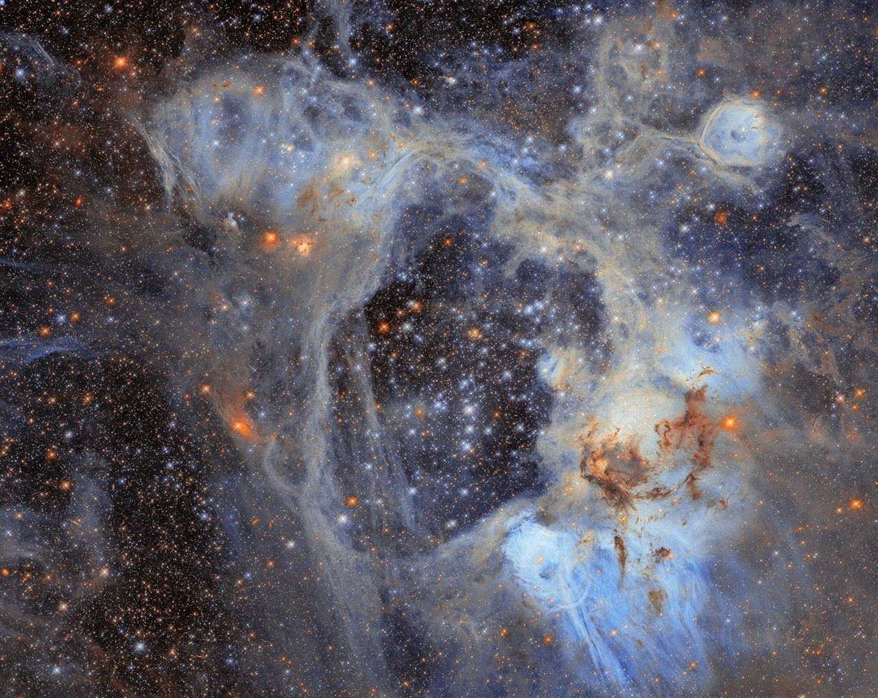 A dense field of stars fills the image, surrounded by wispy clouds of gas and dust. Pale blue and grey nebulosity forms an intricate web across the scene, with darker clouds of dust concentrated towards the lower right. Numerous bright stars appear in shades of blue, white, and orange.