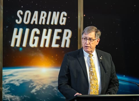 Dan Dumbacher speaks at a podium in front of a backdrop that says “Soaring Higher”
