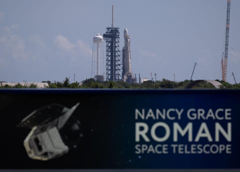 Image shows a banner that reads Nancy Grace Roman Space Telescope. In the background is a rocket standing vertically with a payload fairing on top at the launch pad. Nearby is the mobile launcher, water tower, and a crane in a blue, bright sky.