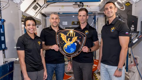 three men and one woman, all wearing black polo shirts, hold a plaque inside a mockup space module