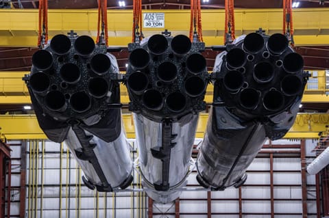 A SpaceX Falcon Heavy rocket is lifted by a yellow crane inside an industrial facility. The 27 black rocket engines stick out of the bottom or three white booster housings, some covered with soot.
