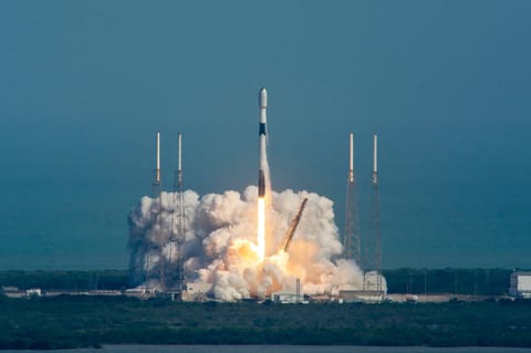 A SpaceX Falcon 9 rocket rises from its launch pad at Cape Canaveral Space Force Station, surrounded by a billowing cloud of white exhaust and a bright orange flame at the base. The rocket climbs vertically against a hazy blue-gray sky, flanked by four lightning rod towers. Launch infrastructure, including a yellow crane arm and concrete support structures, is visible at the bottom of the frame.