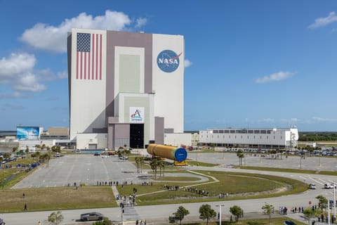 The image shows NASA's Kennedy Space Center iconic Vehicle Assembly Building with an American flag painted on the building along with the NASA and Artemis insignias in a blue sky with some clouds on Tuesday, April 28, 2026.  People gathered to watch the transport of the Artemis III core stage, an orange yellow that will launch during the Artemis III mission. White buildings, grass, and a parking lot can also be seen in the photo. Photo credit: NASA/Glenn Benson
