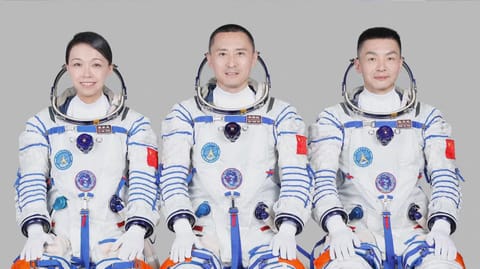 three smiling astronauts &mdash;&nbsp;two men and one woman &mdash; sit wearing white spacesuits