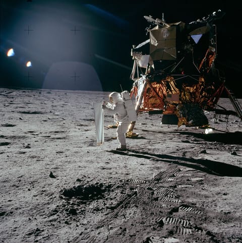 A light brown dust plume, dense at its center and more diffuse toward its edges, extends horizontally across an orange-brown and green landscape. Scattered white clouds cover some of the scene.