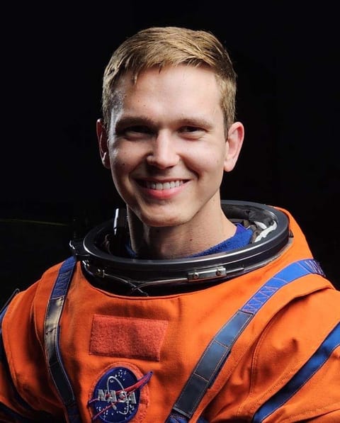 A man poses and smiles wearing an orange spacesuit.