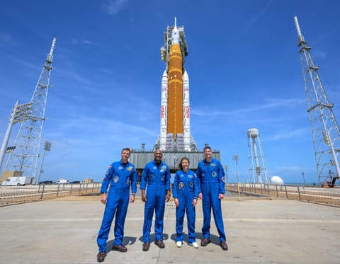 Four NASA Artemis II astronauts at the launch pad with the SLS rocket in the back.