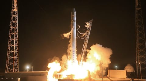 A white SpaceX Falcon 9 rocket launches at night, surrounded by intense orange and yellow flames and large plumes of white smoke. Two tall, metal lattice towers are visible on either side of the ascending rocket. In the background to the right, a white spherical storage tank with the 'SPACEX' logo is illuminated by the launch's glow.
