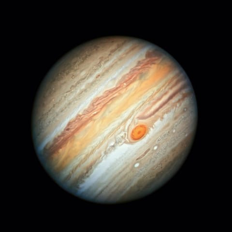 The planet Jupiter, a sphere with horizontal stripes of brown, tan, orange, beige, red, and white appears against a solid black background. The planet’s stripes have varying widths, from very thin to very wide. Within these stripes are mottled, wavy regions, along with several ovals of various sizes. Most of the ovals are whitish, but the largest oval, the Great Red Spot, is orange. The image is taken "sideways" such that Jupiter's stripes run from the upper right to the lower left of the planet
