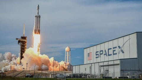 A SpaceX Falcon Heavy rocket blasting off in front of an industrial building with a huge SpaceX logo.