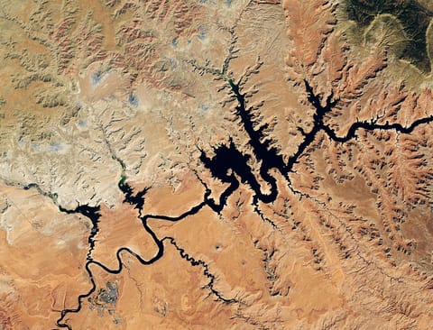 Water in Lake Powell is at record-low levels and looks like a wide river winding through a light orange desert landscape. The Glen Canyon Dam and the town of Page, Arizona, are visible in the lower left.