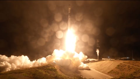 an uncrewed orbital class rocket lifts off into the pitch black sky, lighting its launch pad below with its orange-white plume